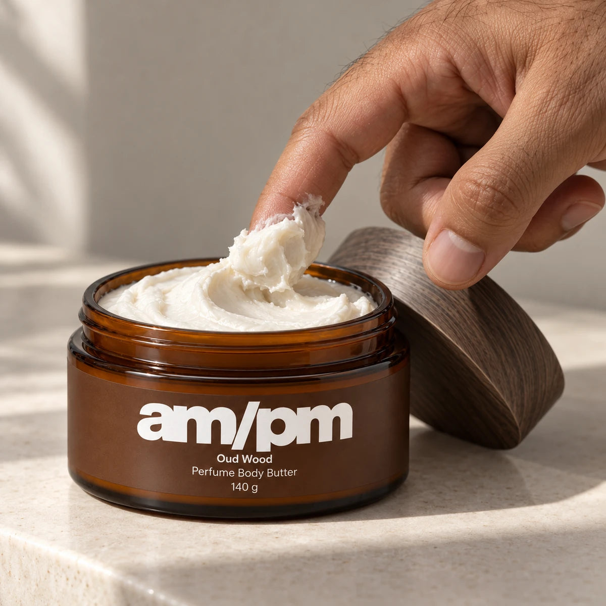 A hand scooping thick ivory body butter from an open amber AM/PM Oud Wood jar.